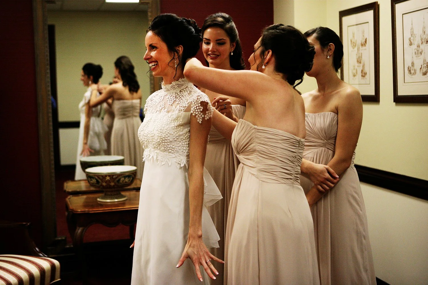 Bridesmaids helping a bride put on her vintage wedding dress in a fitting room with a large mirror behind them. The bride is smiling big.