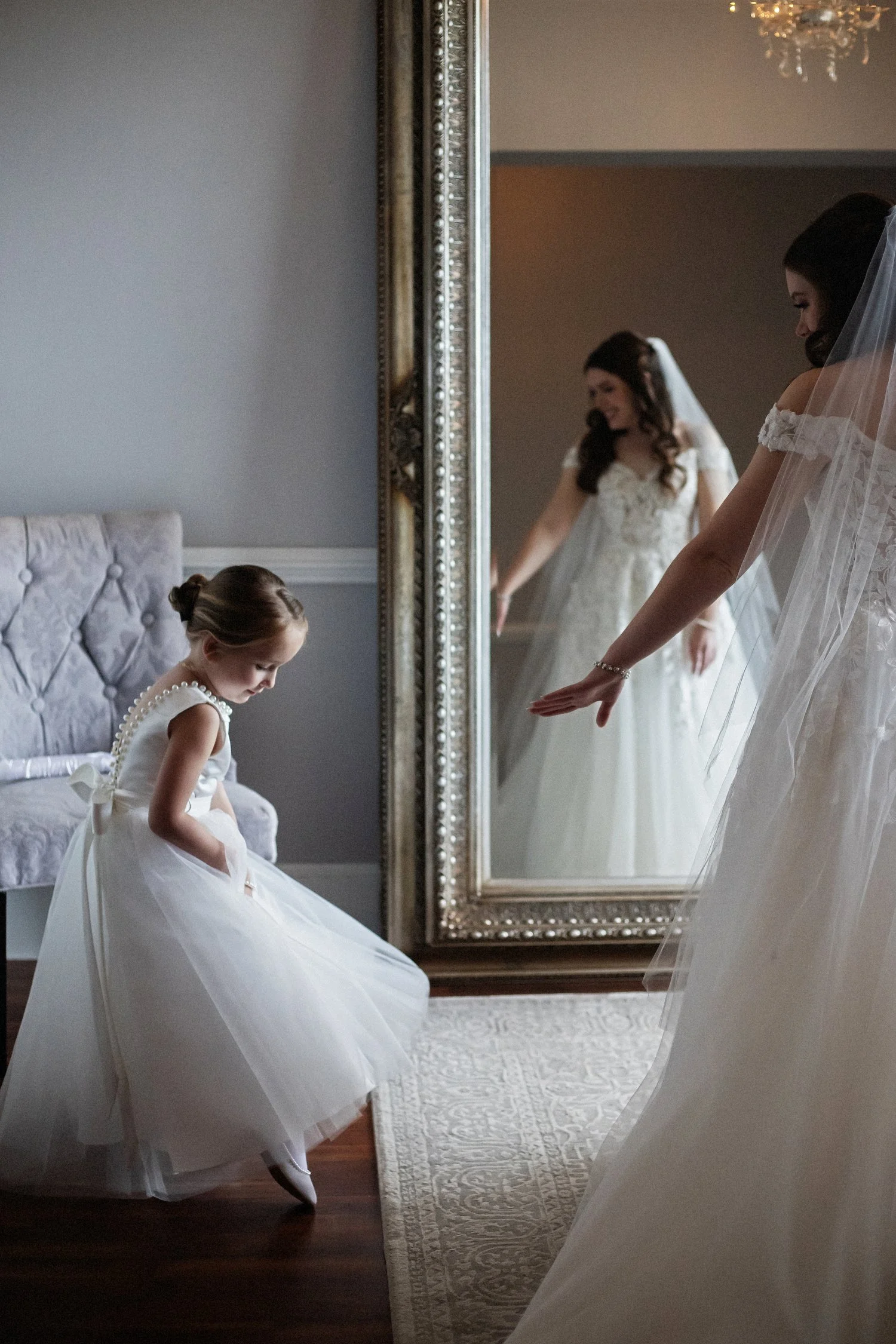 A little girl in a white tulle dress holds her dress up to shoe her shoe to the bride who is next to her reaching her arm out. The bride has on a white gown and veil and her reflection can be seen in a giant silver-framed mirror in front of her.