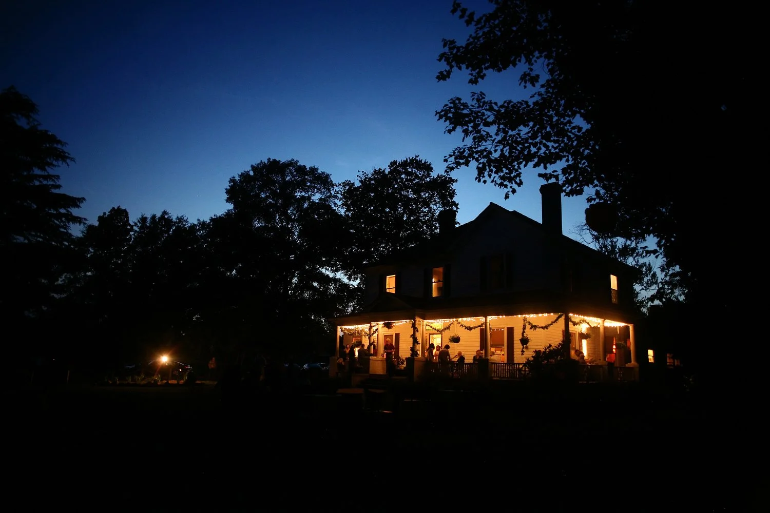 It is dusk, and a huge farmhouse with a wraparound front porch stands in the distance. There are silhouettes of trees all around it, and it is lit with twinkle lights and interior lights in the underneath part of the porch just under the roof. The sk