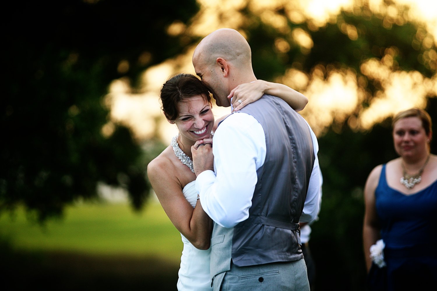 A bride and groom share a joyful dance outdoors during sunset, with a woman in a blue dress watching in the background.