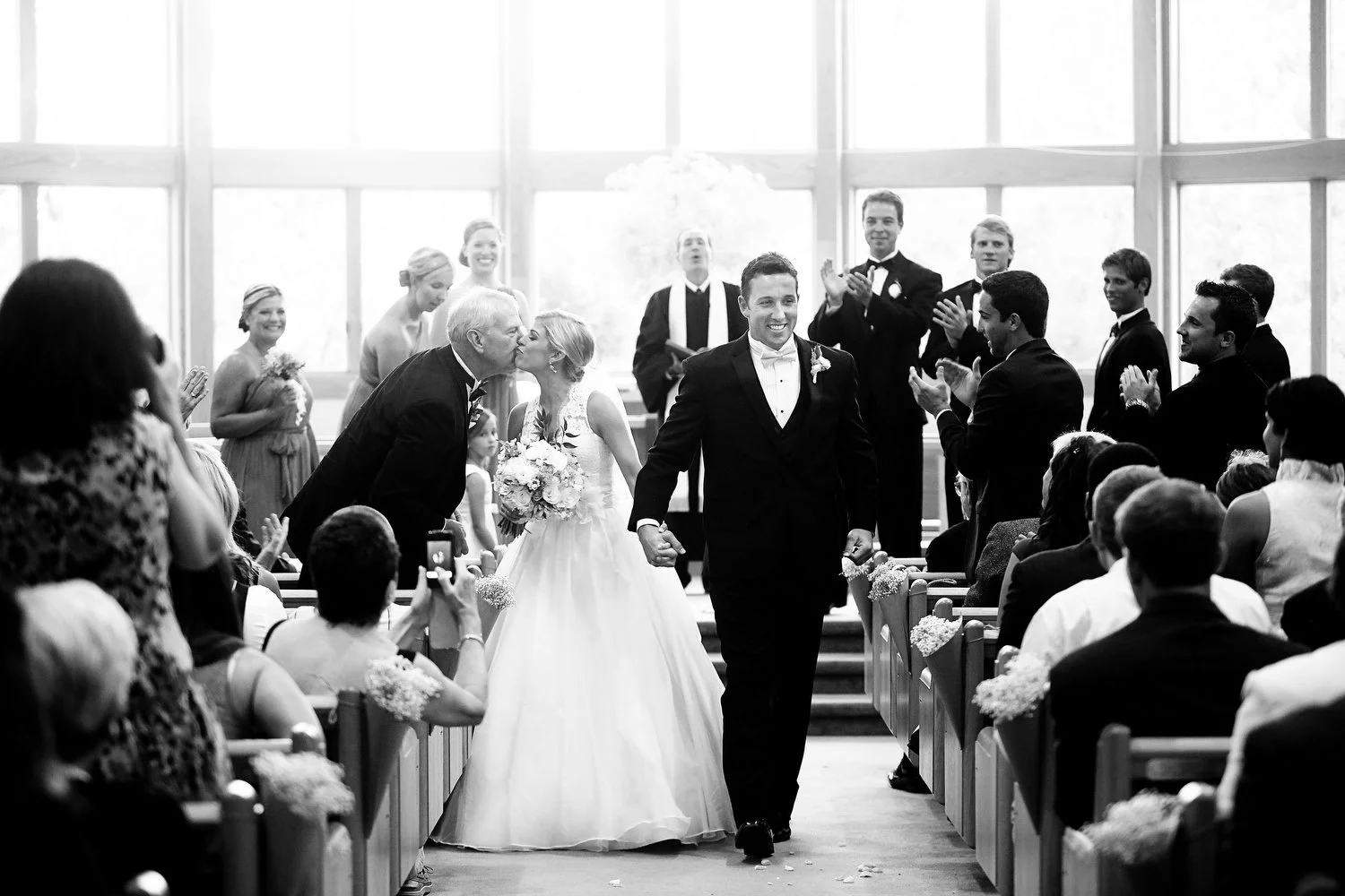 Black and white photo of a newlywed couple walking down the aisle in a church after their wedding ceremony, with family and friends clapping and celebrating. The bride leans over to kiss her father. They have huge floor length windows behind them.