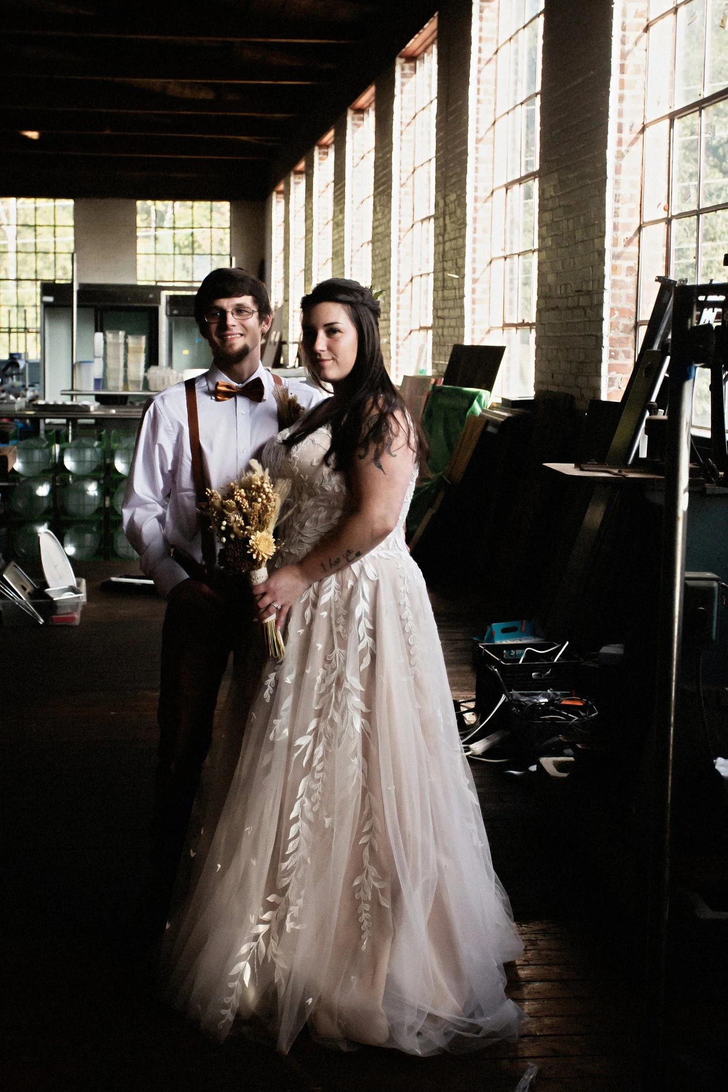 A bride and groom stand in an old building with huge windows all around and various equipment and materials around them. She has on a vintage white lacy dress. He has on a white dress shirt, suspenders and a bowtie. 