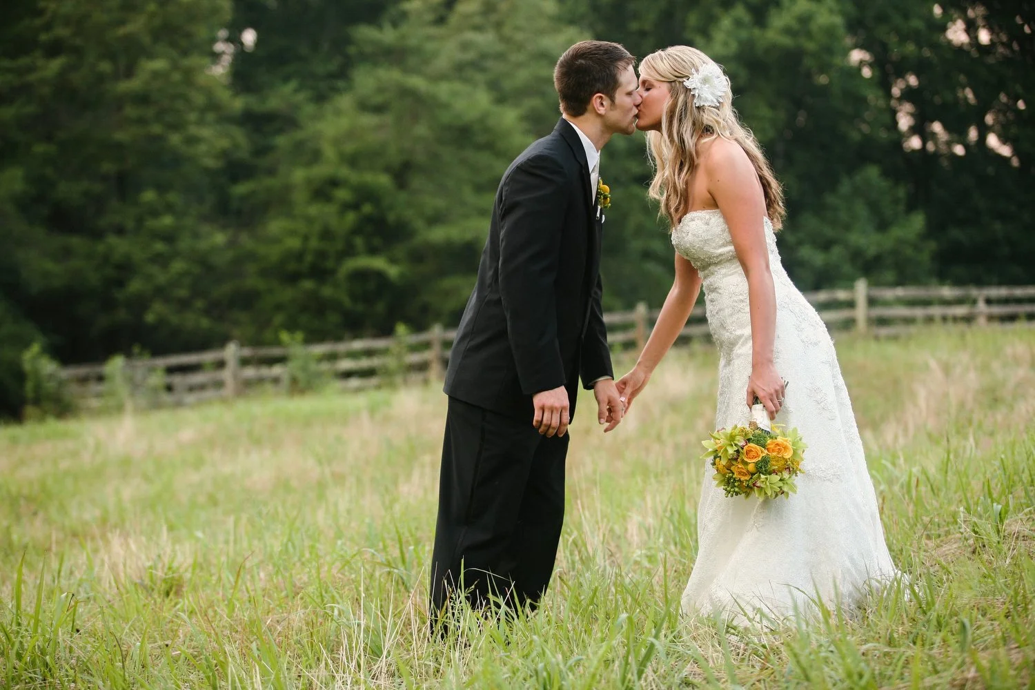 A bride and groom hold hands and lean in to kiss in a grassy meadow with lush green trees and a wooden farm fence in the distance. He has on a black suit. She has on a strapless white dress with a white flower piece in her long blond hair. She clutch