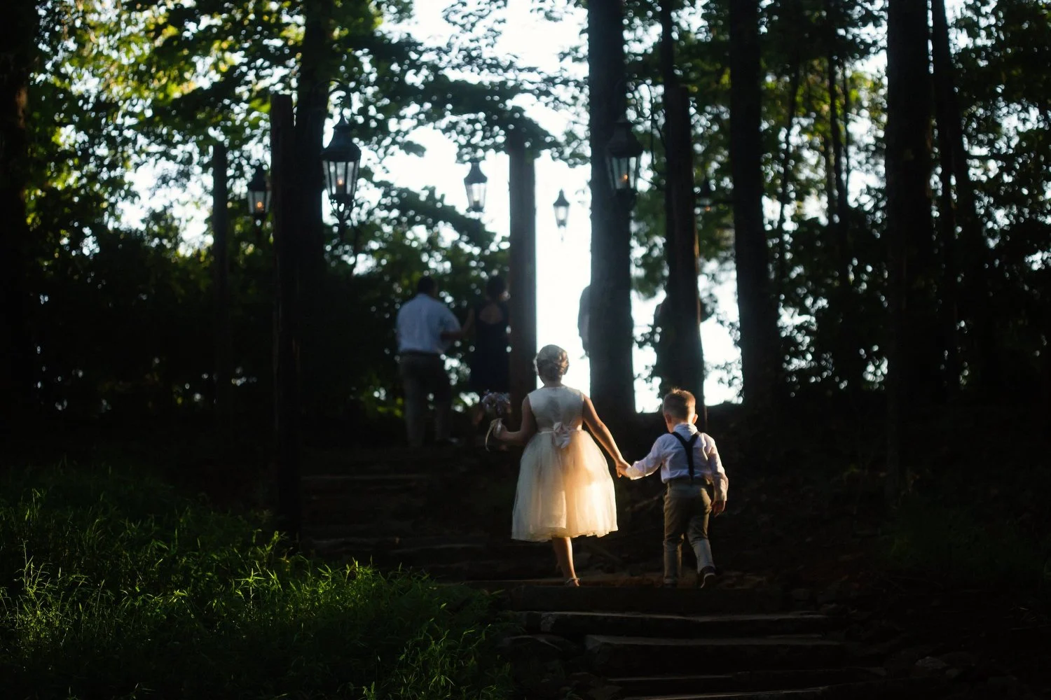 A young girl in a sheer dress holding hands with a boy in suspenders and dress shirt, walking up steps through a forested area with lanterns, silhouetted by the late-day sunlight.
