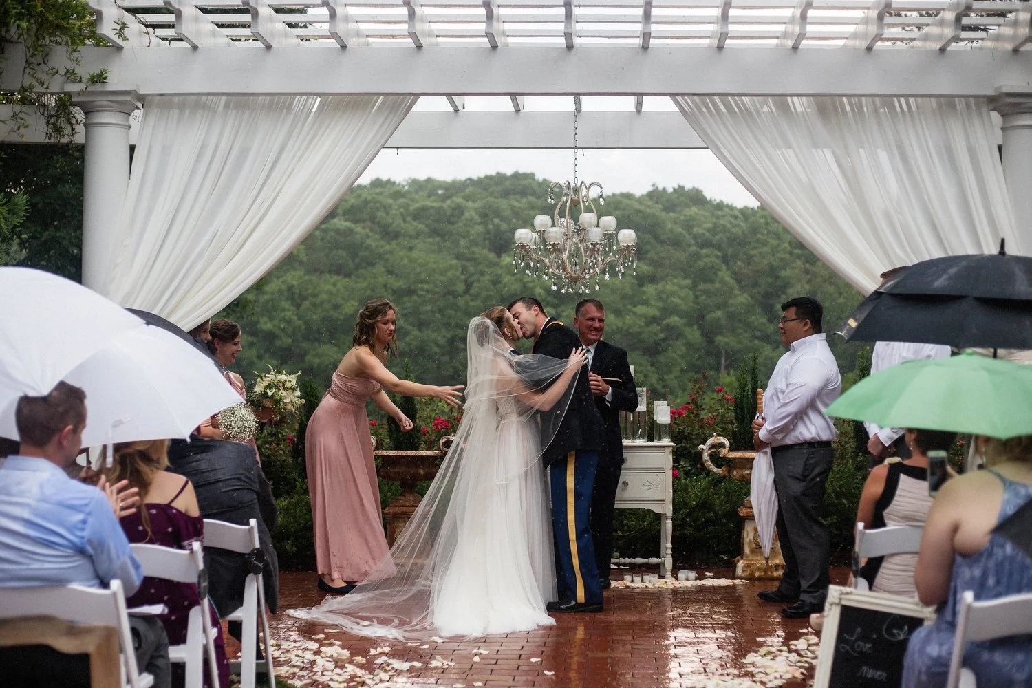 A wedding ceremony taking place outdoors under a white canopy, with a bride and groom kissing, surrounded by guests holding umbrellas in rain.