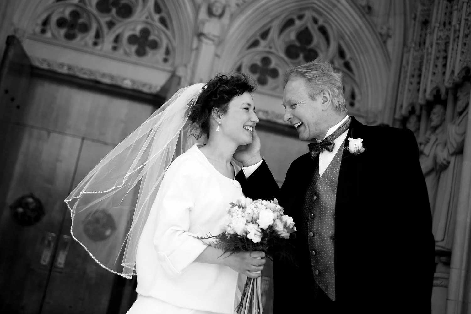 A bride and an older man, possibly her father, sharing a happy moment during a wedding ceremony just outside a church. The bride is holding a bouquet of flowers, wearing a veil, and smiling as the man gently touches her face. They are standing just o