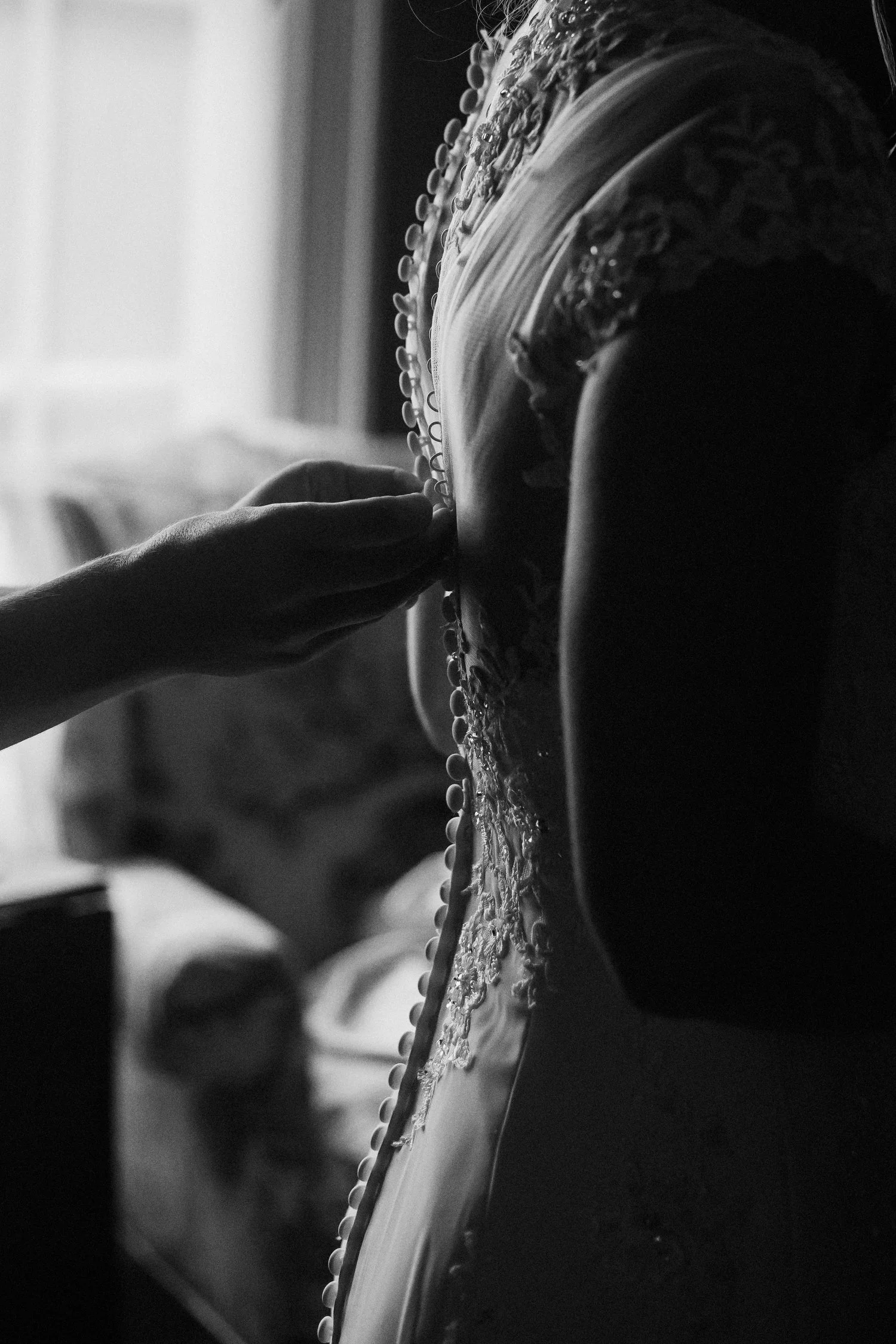 Hands work to fasten the buttons on the back of a wedding dress on a bride with intricate detail, in black and white, with natural light coming through a window in the background.