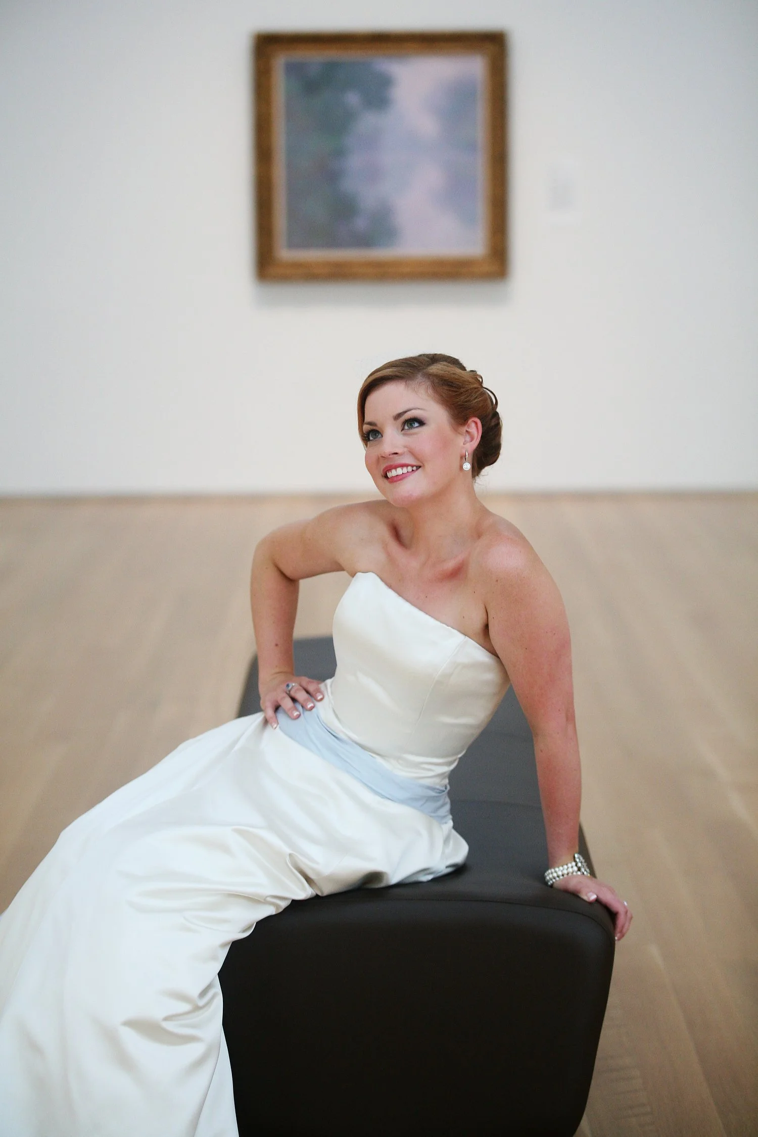 A bride in a strapless white dress with a decorative belt poses on a soft rectangular bench with one hand on her hip. She is in an art gallery with light wooden floors and a painting is framed on the wall in the distance behind her.