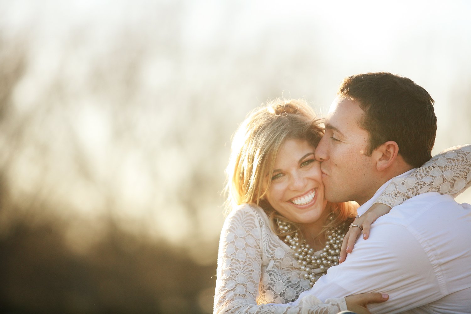 A couple embraces in a tight image where they are in the front lower right corner of the frame. They are slightly backlit by the late sunset glow behind them. Blurry trees or a wild field is visible in the far distance behind them. She is wearing a l