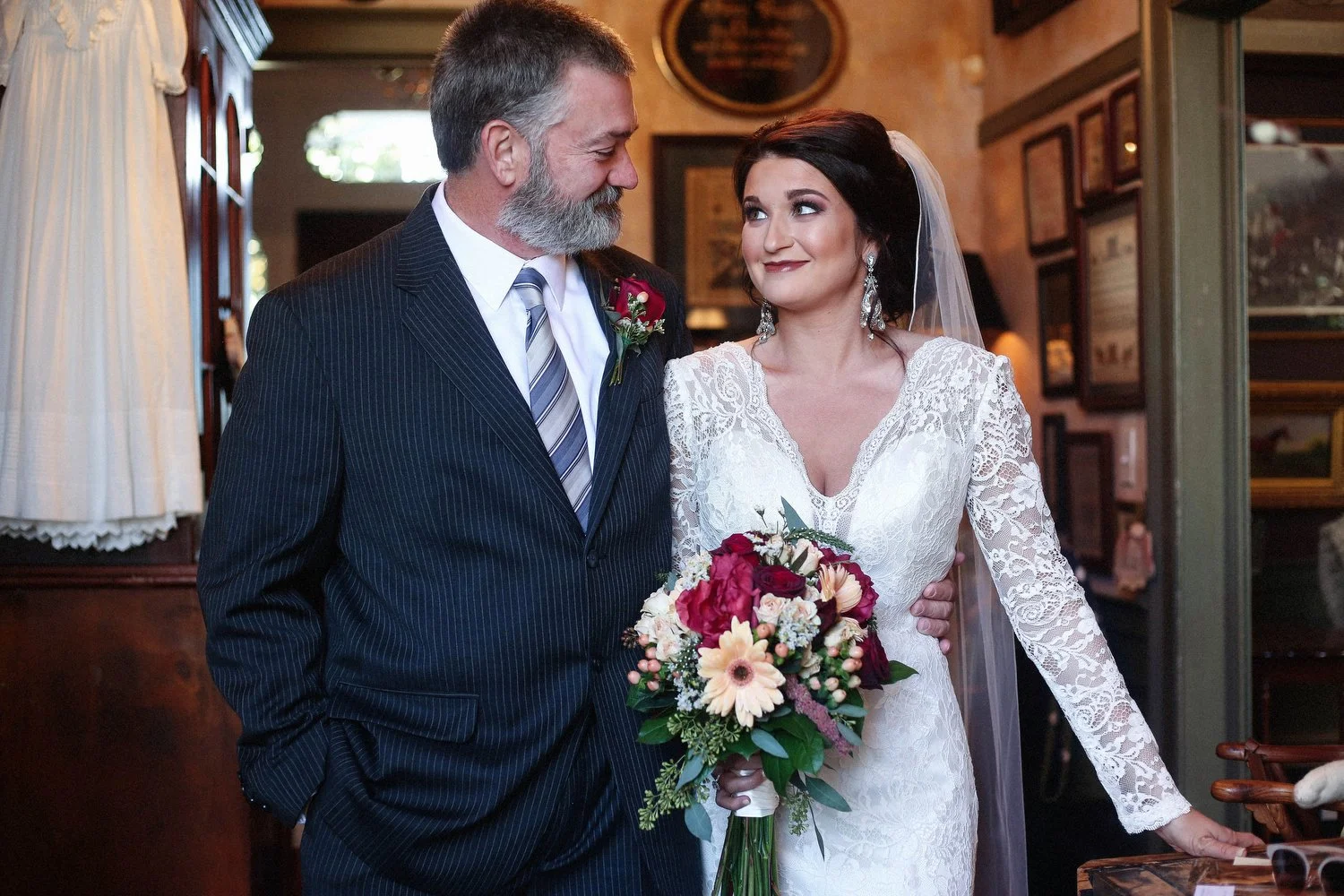 A bride and an older man, likely her father, in a doorway. The bride hold a bouquet of flowers and wears a lace wedding dress with a veil. She looks at him with a smile. The man is wearing a dark pinstriped suit with a white shirt and a striped tie.