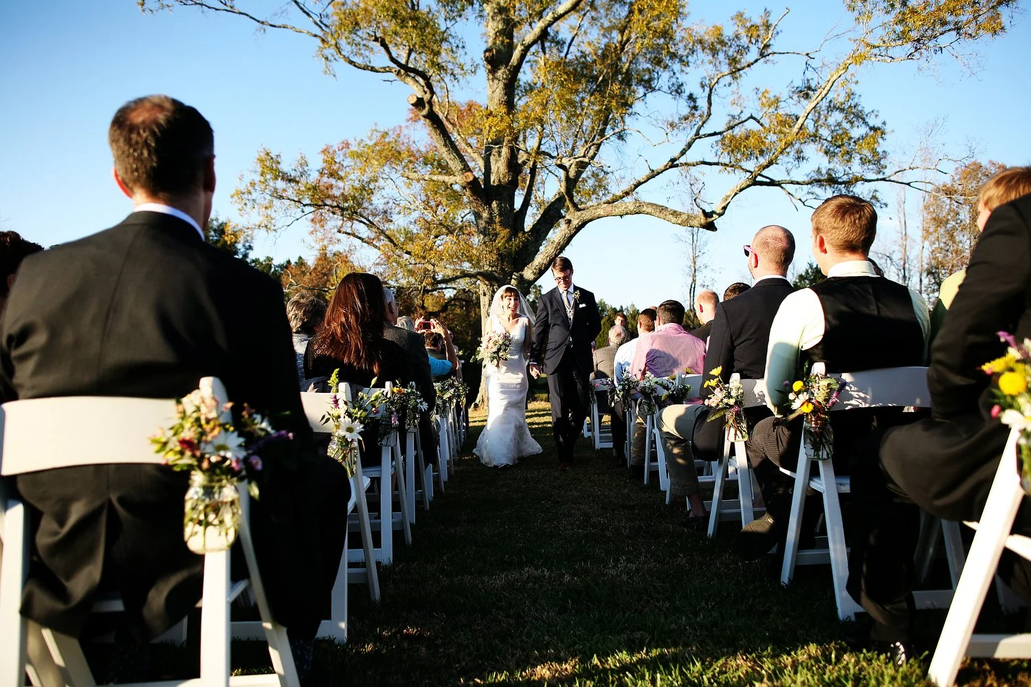 Outdoor wedding ceremony with guests seated on white chairs, bride and groom walking down aisle, under a large tree with fall foliage, bright sunny day.
