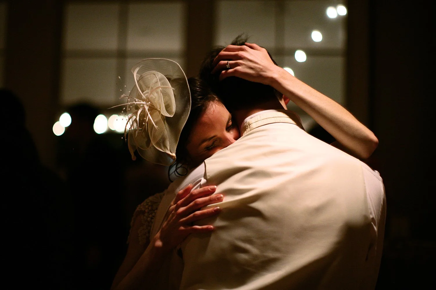 Close photo of a bride and groom embracing in a dimly-lit reception. He is seen from behind in a white dress shirt, and she leans her face on his shoulder and clasps his back and the back of his head with her arms. She has a fascinator on her head.