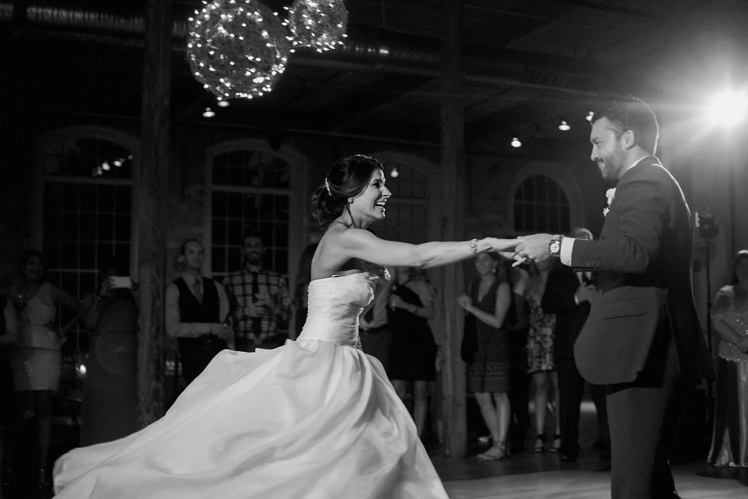A bride and groom sharing a dance at their wedding reception, with guests watching in the background, during an indoor celebration with dim lighting and decorative hanging lights. Her arm is outstretched as he prepares to spin her.