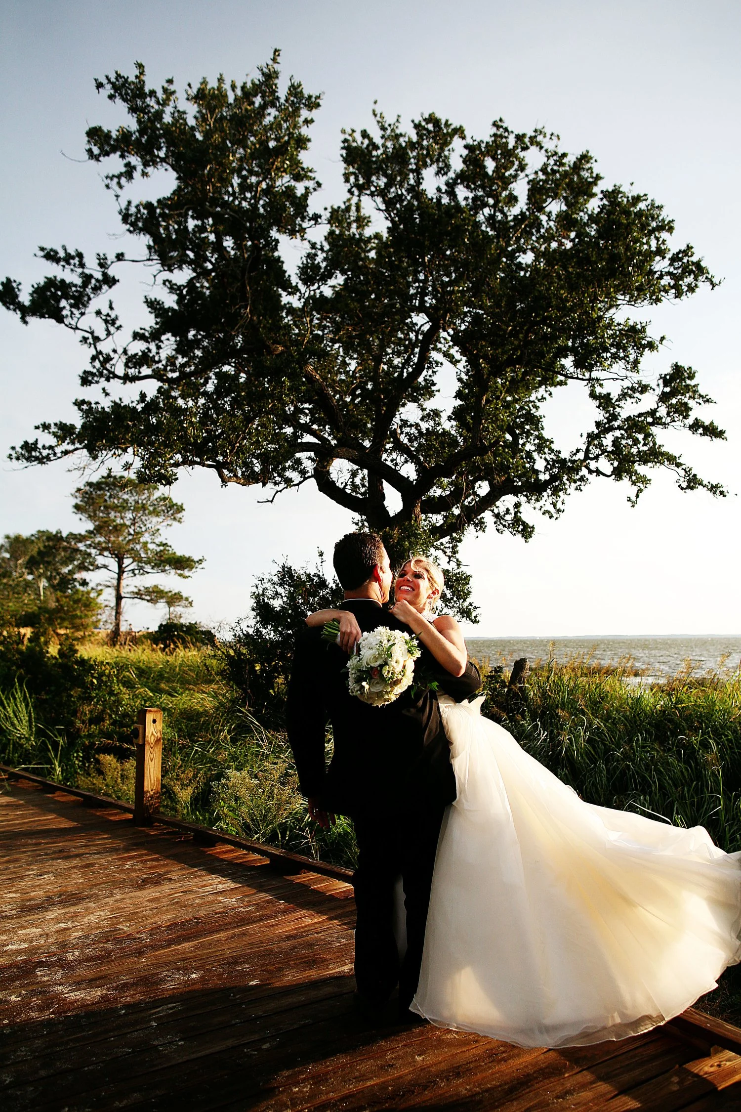 A couple in wedding attire, the woman in a white wedding gown holding a bouquet, embracing and smiling for a photo outdoors on a wooden path, with grass and a large tree, near a body of water in evening sunlight.