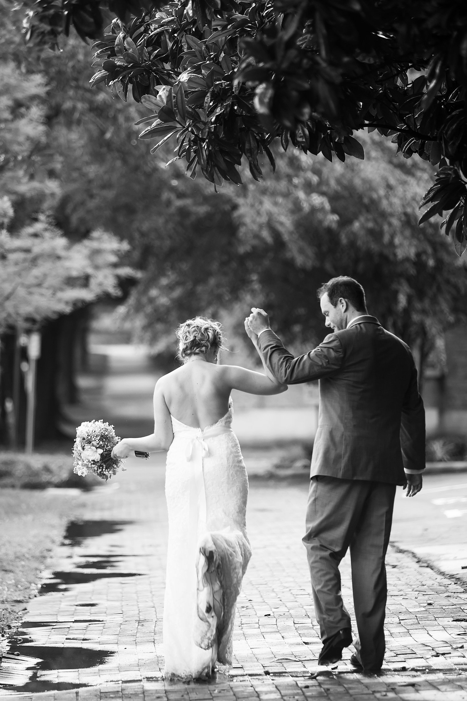 A black and white photo of a bride and groom walking hand in hand on a brick pathway, the bride holding a bouquet of flowers and wearing a strapless dress, while the groom wears a suit. They are surrounded by trees.