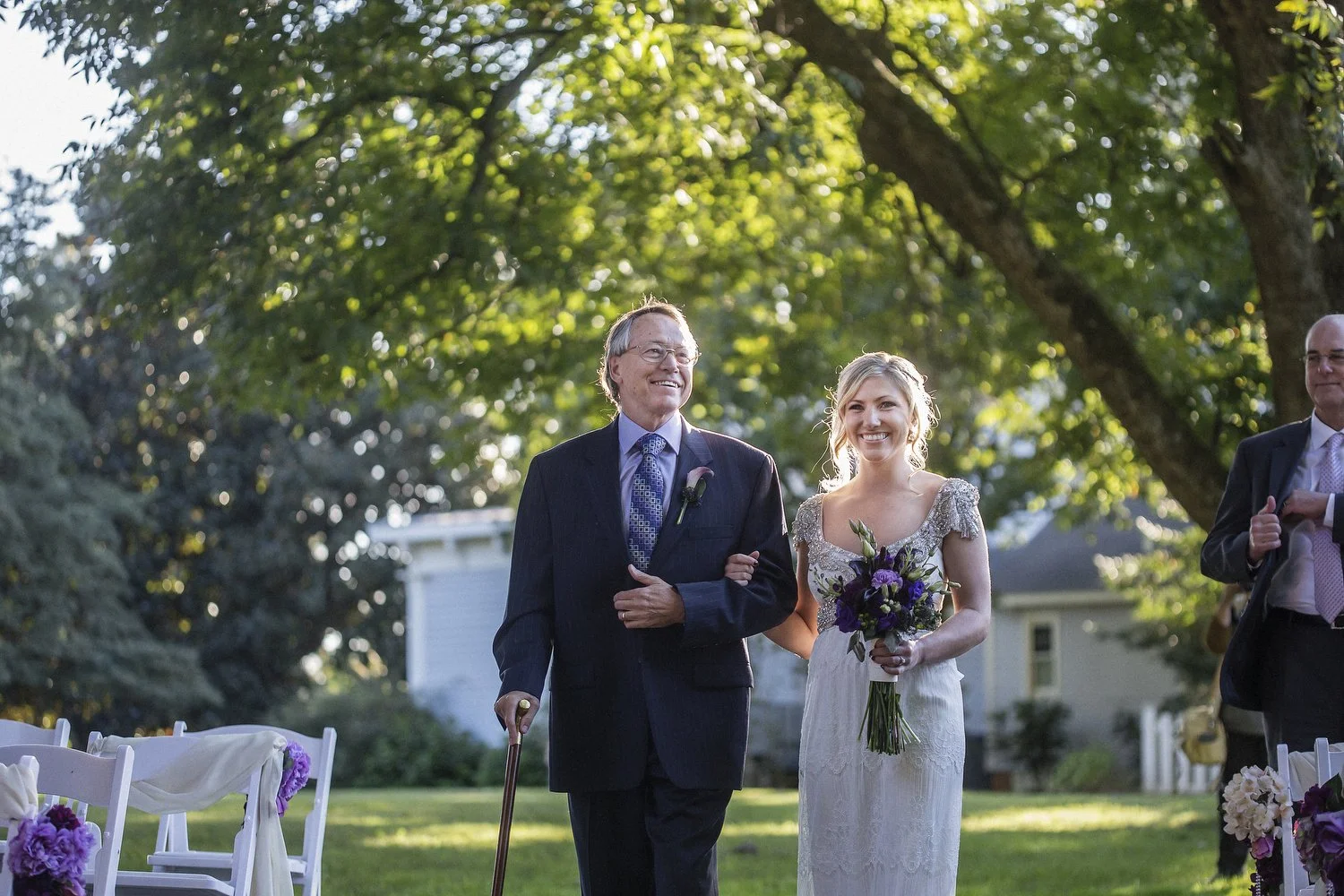 Bride in vintage white wedding dress with decorative sleeves holding a bouquet of purple flowers walks arm-in-arm with her father down the aisle. He is wearing a black suit and glasses and using a cane. There are white chairs and large trees around.