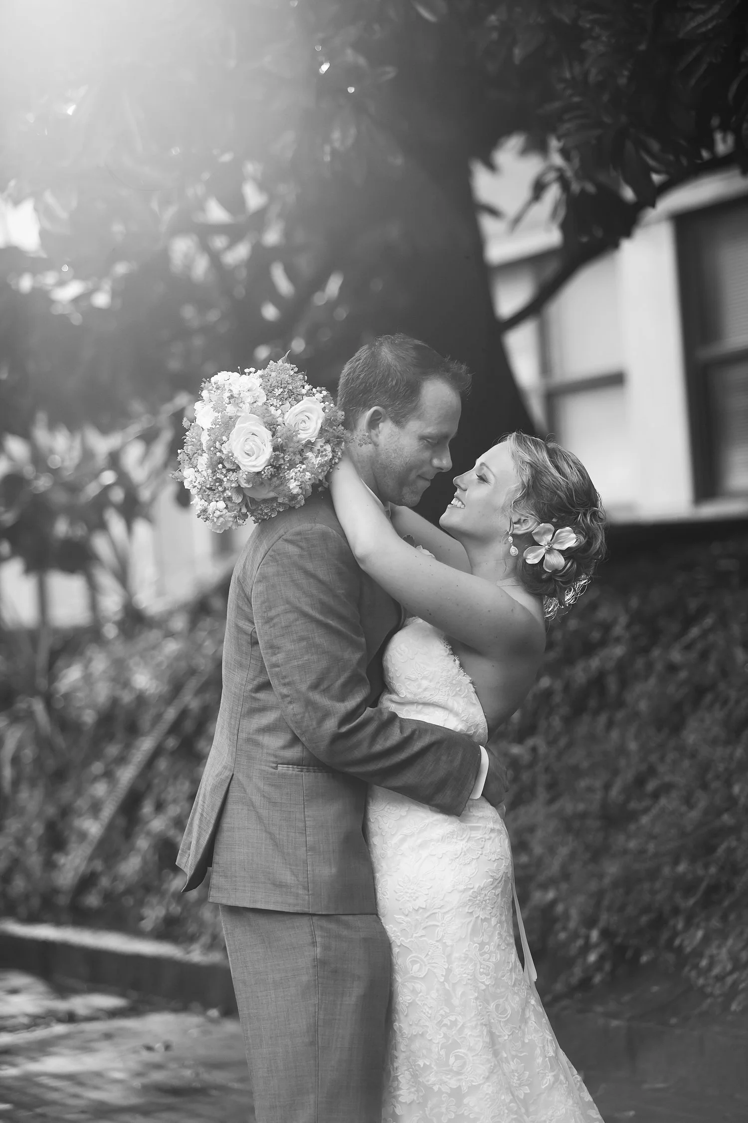 Black and white photo of a bride and groom embracing outdoors, with trees and a building in the background. The bride is holding a bouquet of flowers and is smiling at the groom.