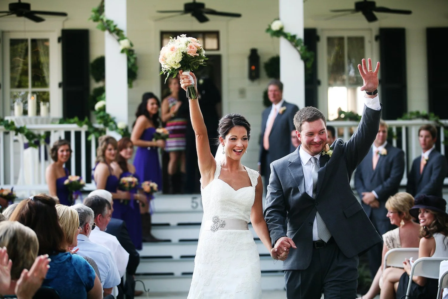 A bride and groom celebrating their wedding outdoors, with the bride holding a bouquet and the groom waving to guests as they walk down the aisle after their ceremony.