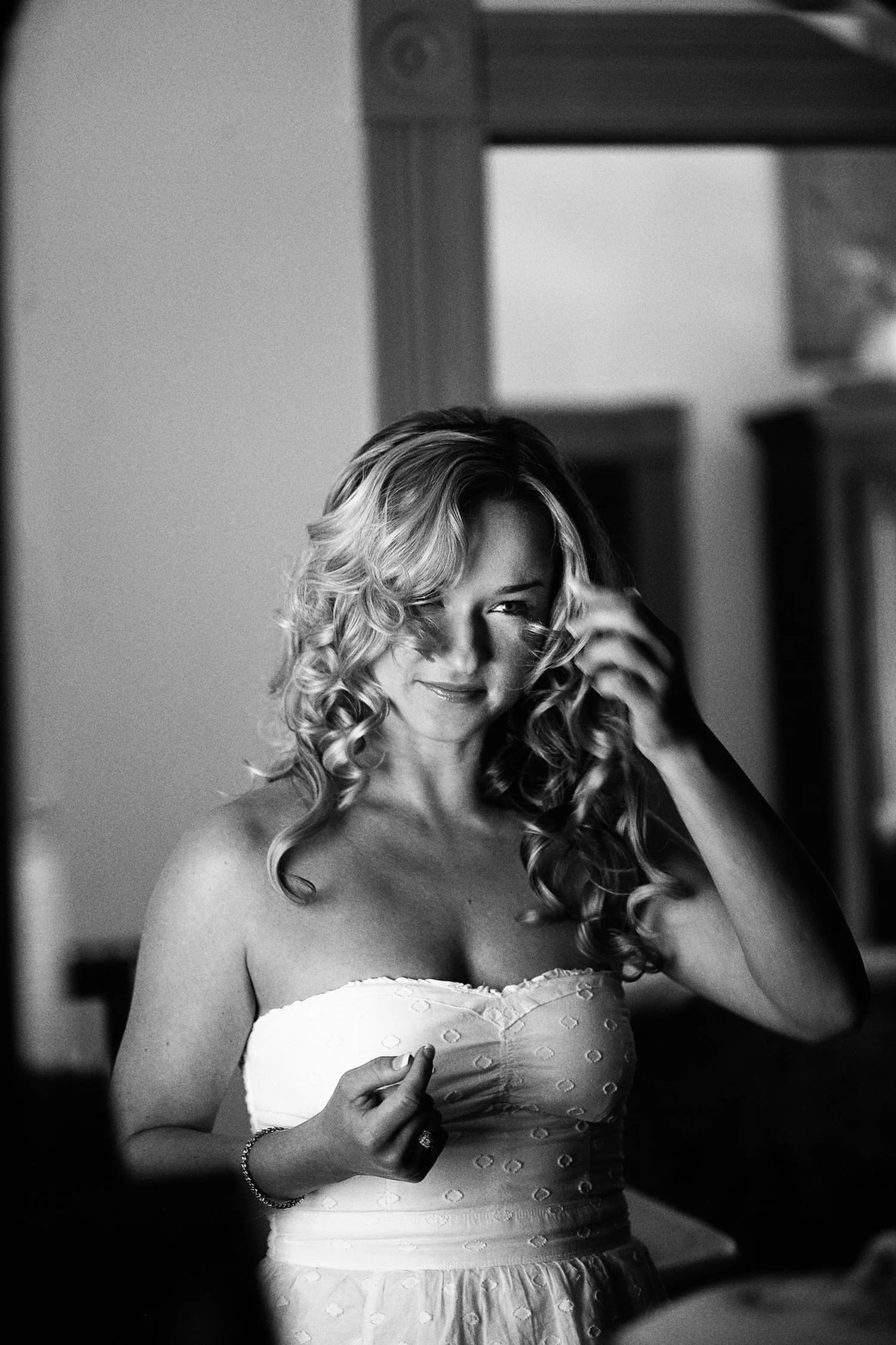 Black and white image of a woman with curly blonde hair smiling and adjusting her hair, wearing a strapless white dress.