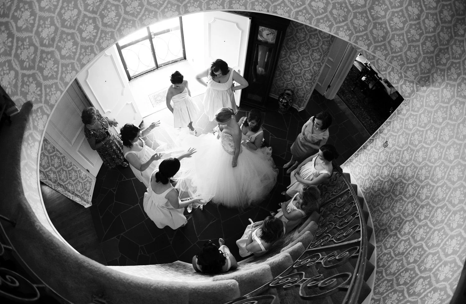 A black and white photo of a bride in white tulle surrounded by bridesmaids and family members inside a house with decorative wallpaper, taken from the top of a curved staircase with a wrought-iron rail.