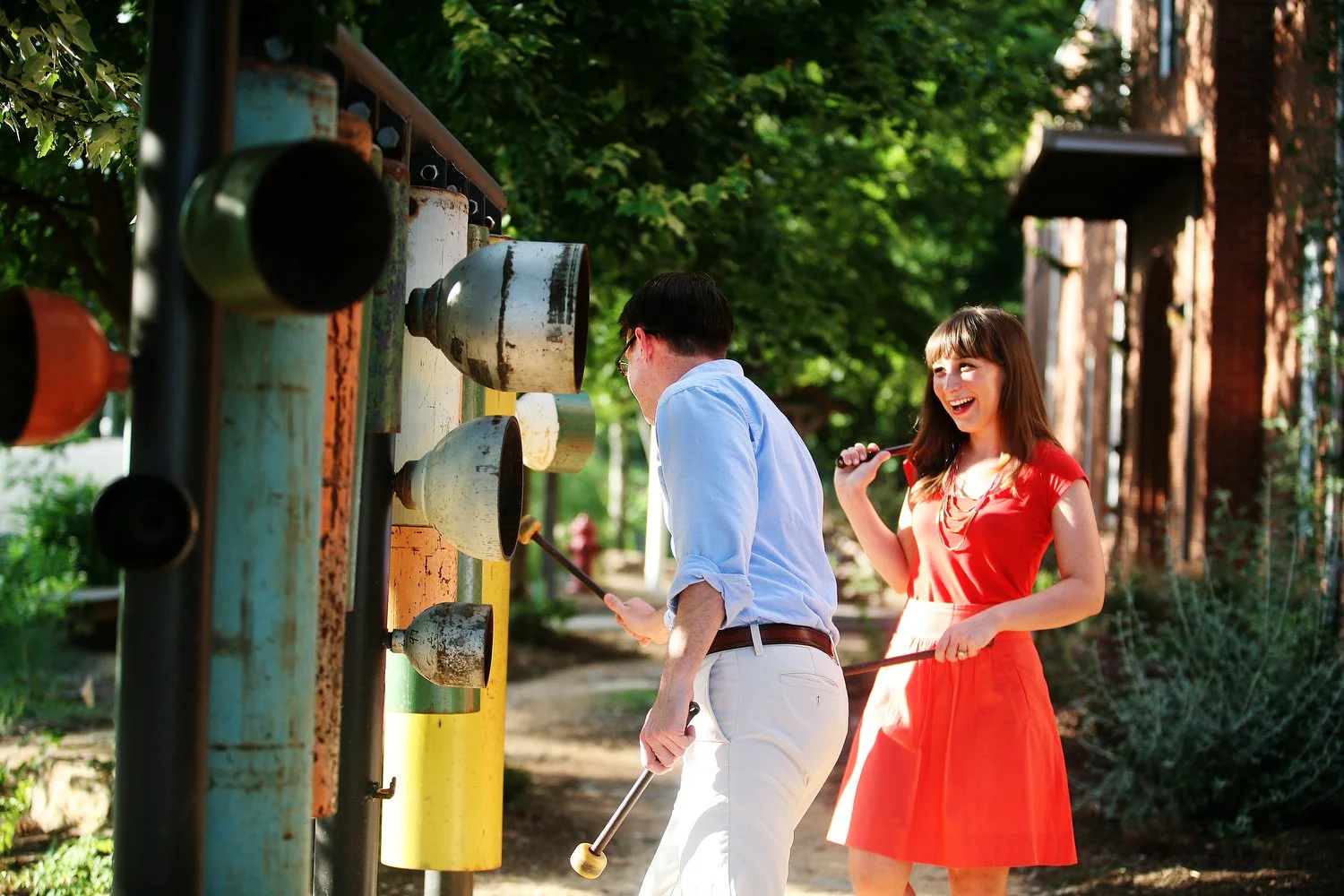 A couple playfully interacts with a metal stoplight looking structure. They have rubber mallets in their hands and appear to be laughing as they step back from playing music on the metal structure. She has on a short red dress and her dark hair is st