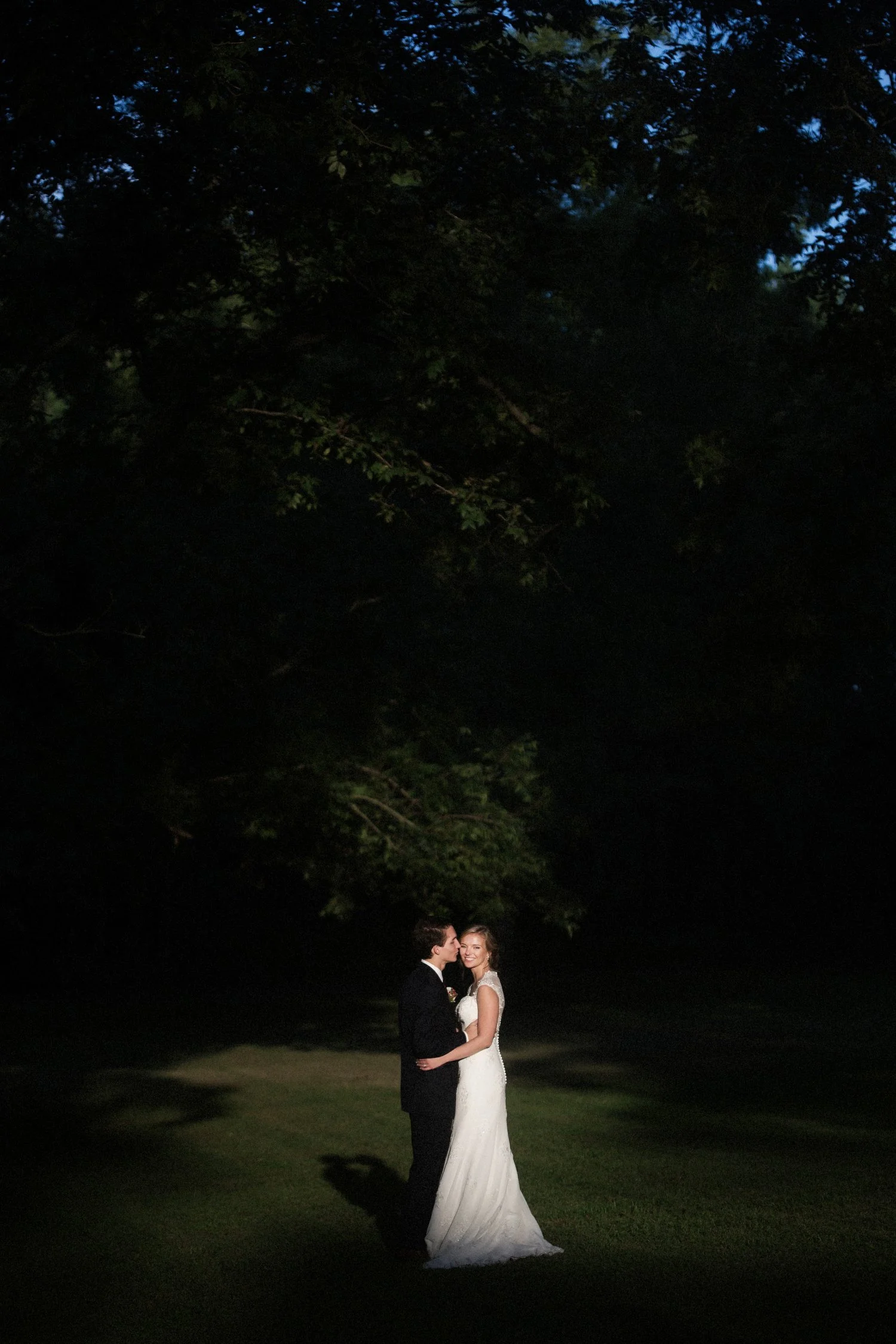 A bride and groom embracing outdoors in dramatic light with dark green trees surrounding them, illuminated by the sunlight.