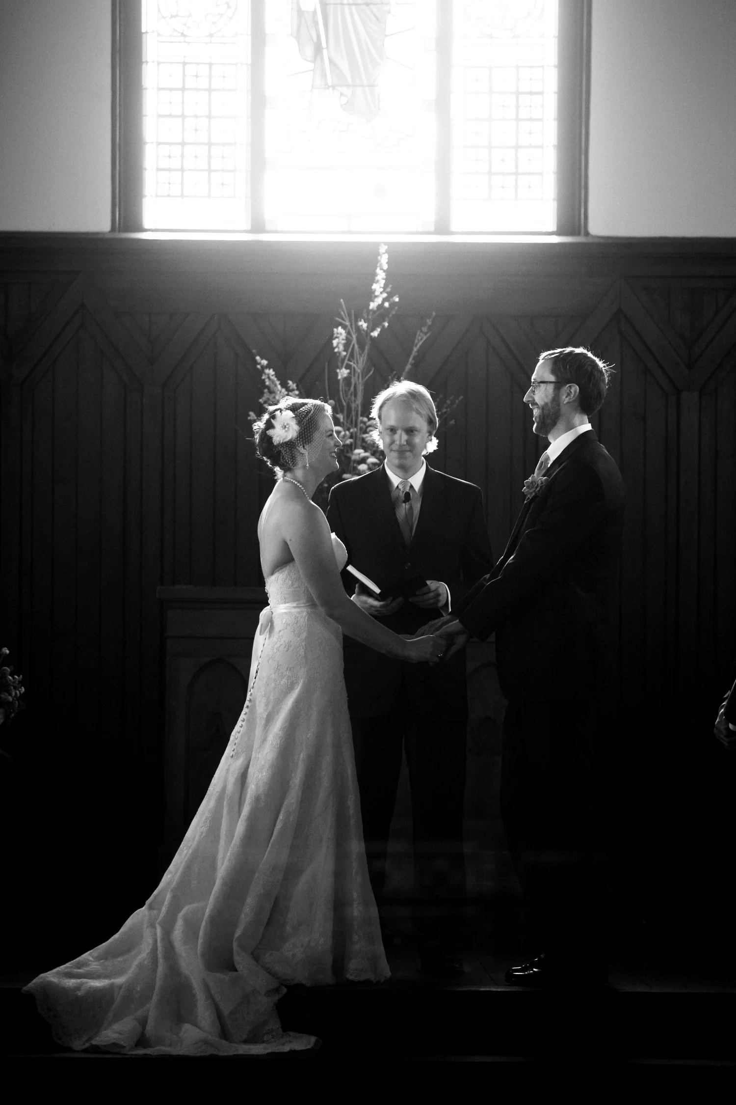 A black and white photograph of a wedding ceremony with a bride and groom holding hands, standing in front of an officiant inside a church. The bride is wearing a strapless gown with a long train, and the groom is dressed in a dark suit with a white 
