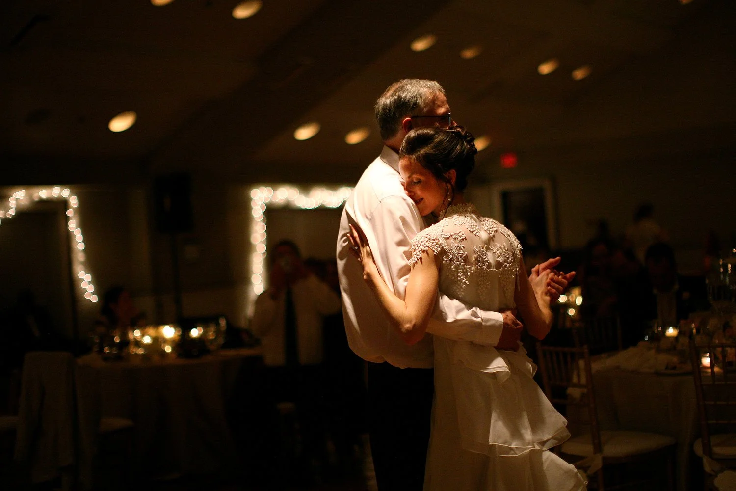 A bride hugs her father tightly as they dance closely at a wedding reception, with guests seated in the background, dim lighting, and soft candles on the tables.