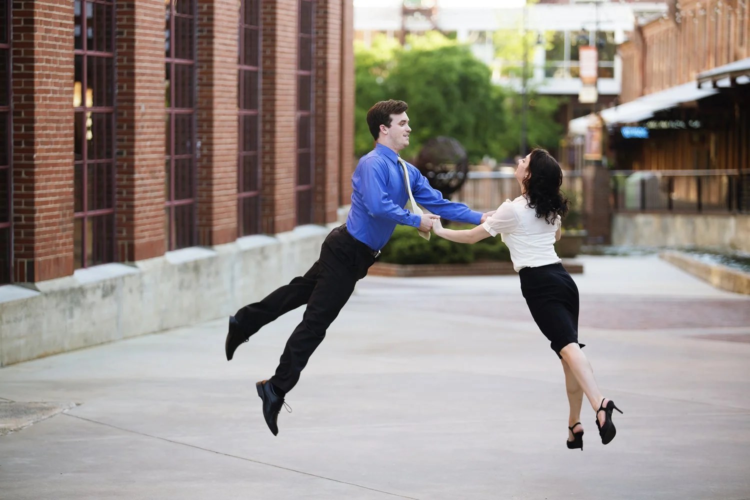 A couple dances on a concrete path in between brick buildings with green trees in the distance behind them. He has on a blue dress shirt, black dress pants and a tie. She has on a white dress shirt, a black mini skirt and black high heels. They are h