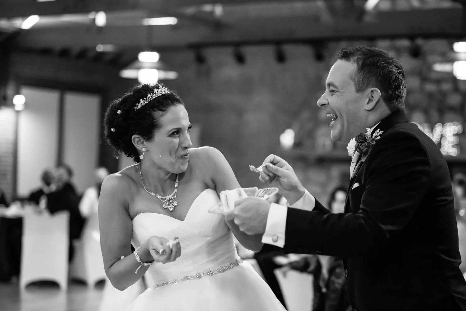 Black and white photo of a bride and groom smiling and sharing cake at their wedding reception, with guests in the background.