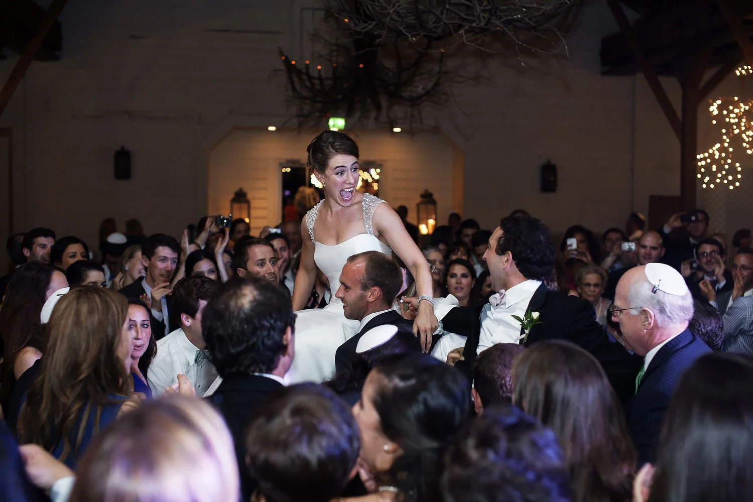 A woman in a wedding dress being lifted in a chair and celebrated at a wedding reception with many guests surrounding her in the Jewish hora tradition.