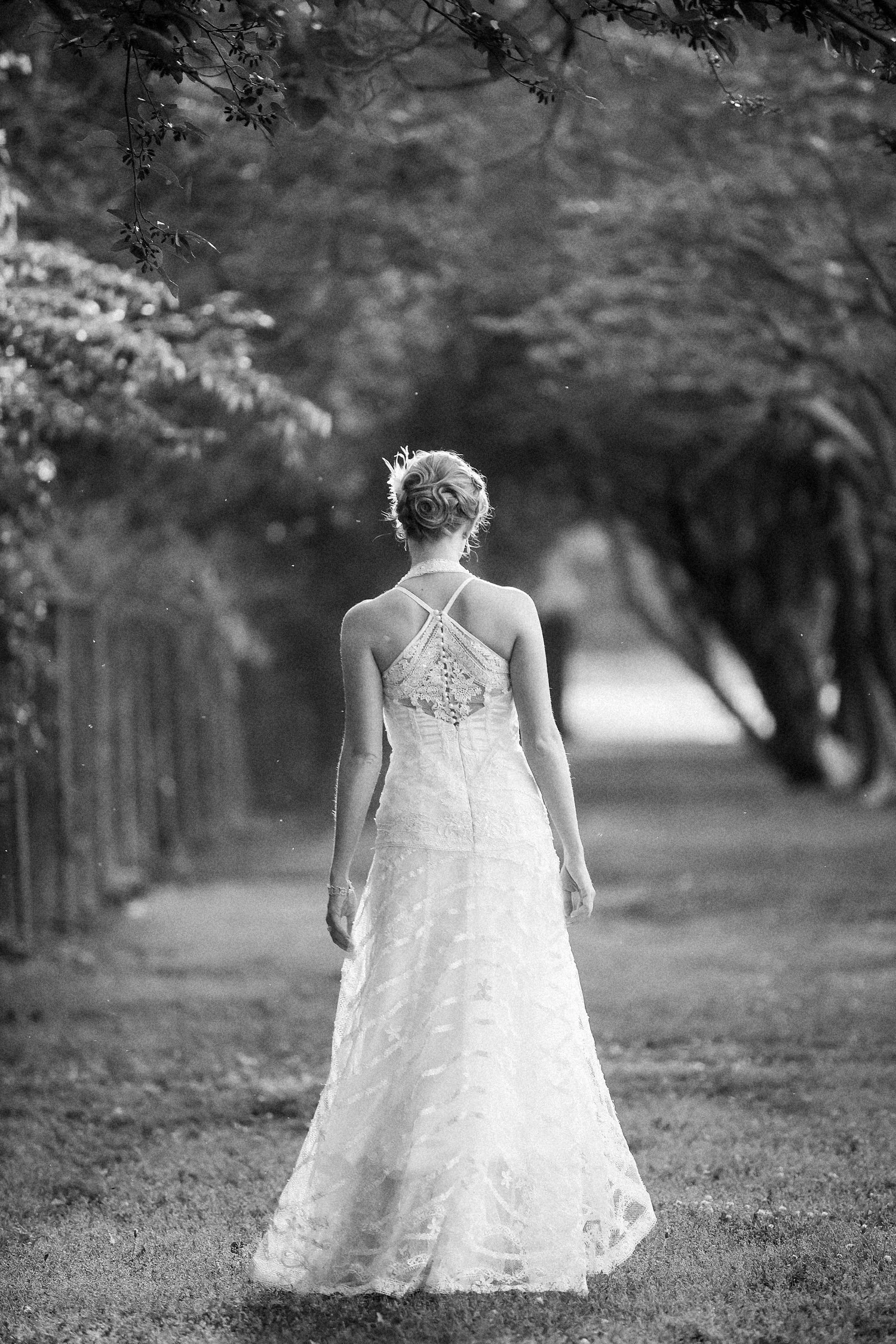 Black and white photograph of a woman in a wedding dress walking away on a tree-lined path.