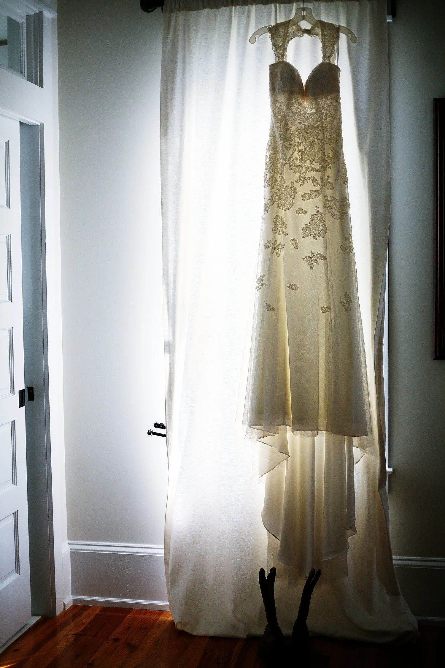 Sheer cream wedding dress with decorative appliqués on it hangs from a floor-length window with sheer lace curtains behind it. There are cowboy boots on the wooden floor underneath the dress.