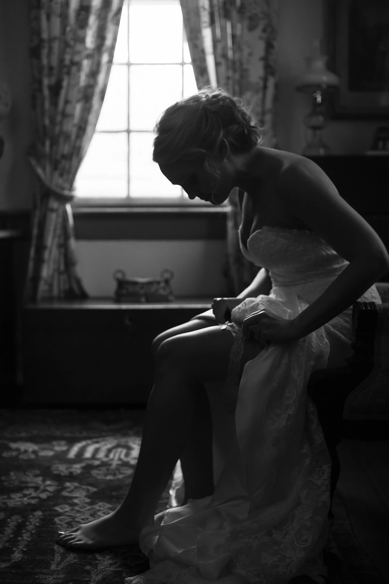 Black and white silhouette of a bride sitting down and leaning over while hiking her dress up to slide her garter up on her thigh. There is a window with curtains pulled to either side behind her. 