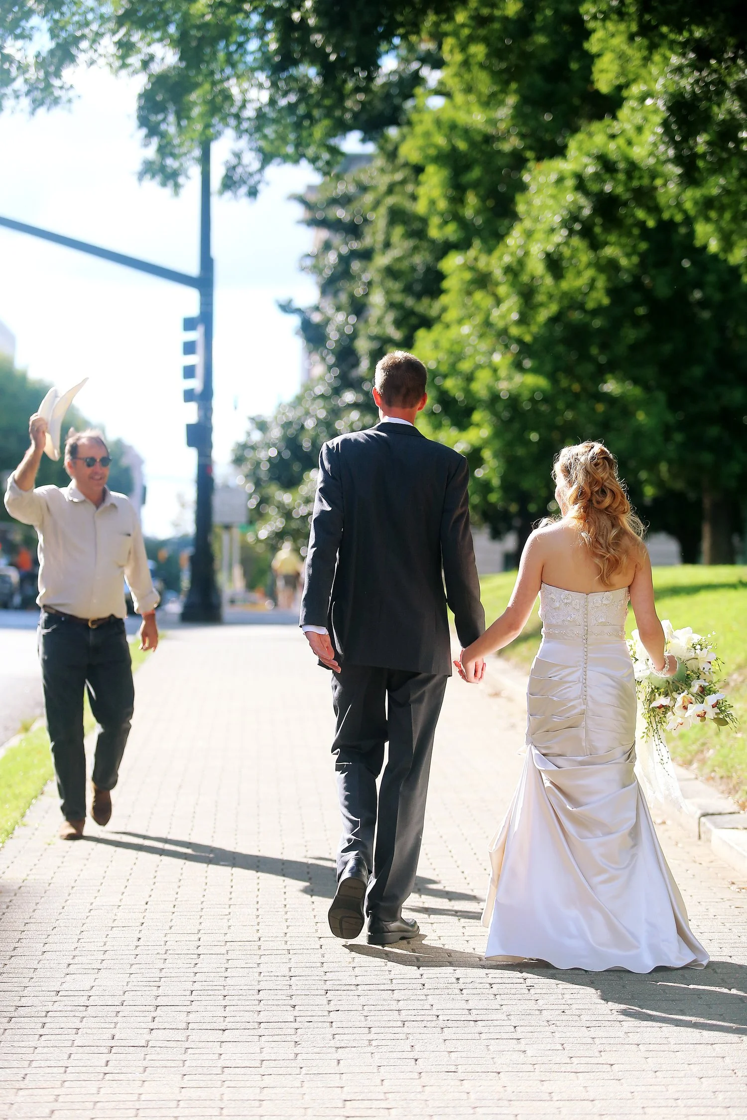A bride and groom walking hand in hand on a sidewalk, with a man waving and holding out his cowboy hat as he walks toward them, against a backdrop of green trees and a sunny sky. The bride has on a strapless white dress and her blond hair is styled i