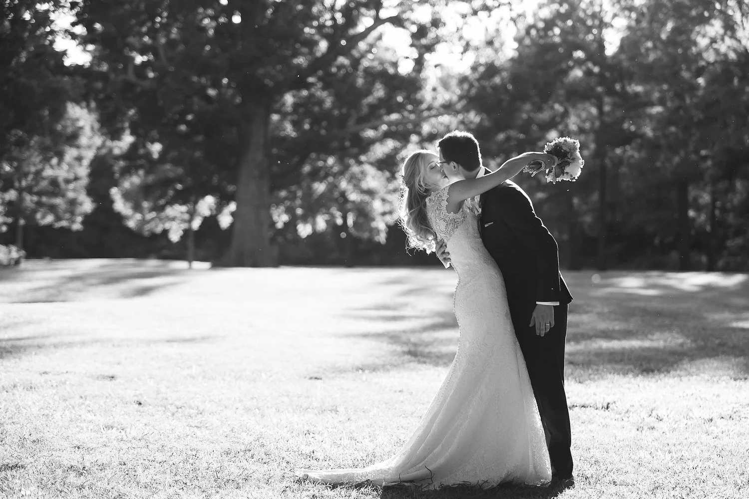 A black and white photo of a bride and groom kissing outdoors on a grassy area, with the bride holding a bouquet and wearing a lace wedding gown, and the groom dressed in a dark suit.
