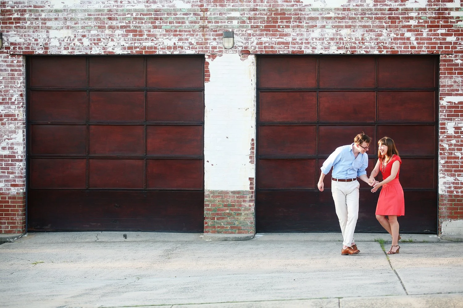 Two sets of large square-shaped doors looking like garage doors are framed by old red and white bricks. The doors are dark brown and look to be made of metal. In front of the door on the right stands a couple small in the frame. She is wearing a red 