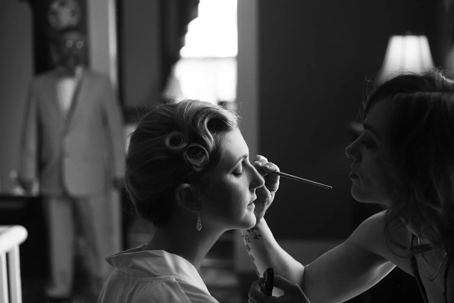 Black and white photo of a bride with vintage hair being styled by a makeup artist in a room with a window and a lamp. Her father stands in the background in a light-colored suit.