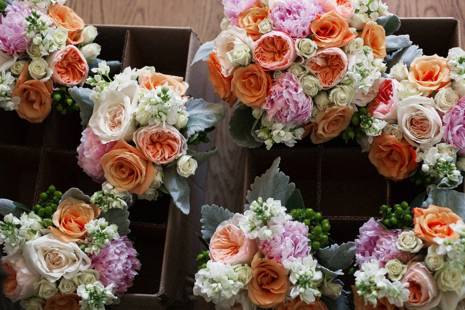 Seven bridal bouquets are photographed from above. They are in cardboard boxes with square sections. The bouquets are made up of orange, peach, pink and white flowers including roses, peonies and ranunculus. 