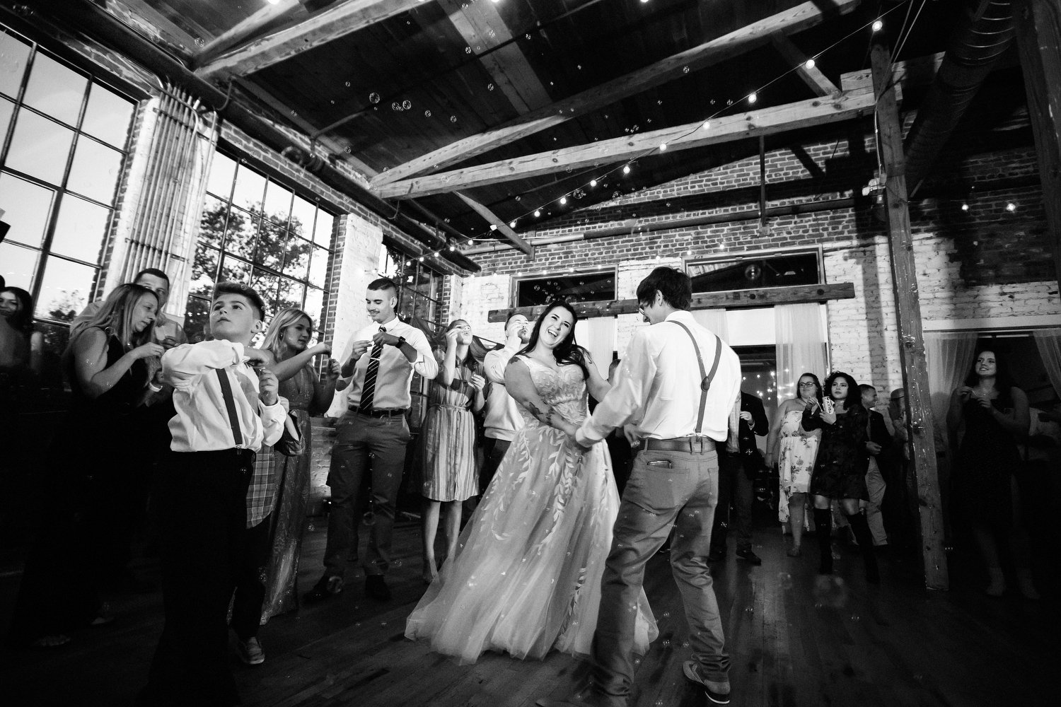  Black and white wedding reception image taken from a low perspective. The bride and groom are dancing. She has on a floor-length white dress and holds his hands while leaning to the side and smiling. He is seen from the back in tan dress pants, a wh
