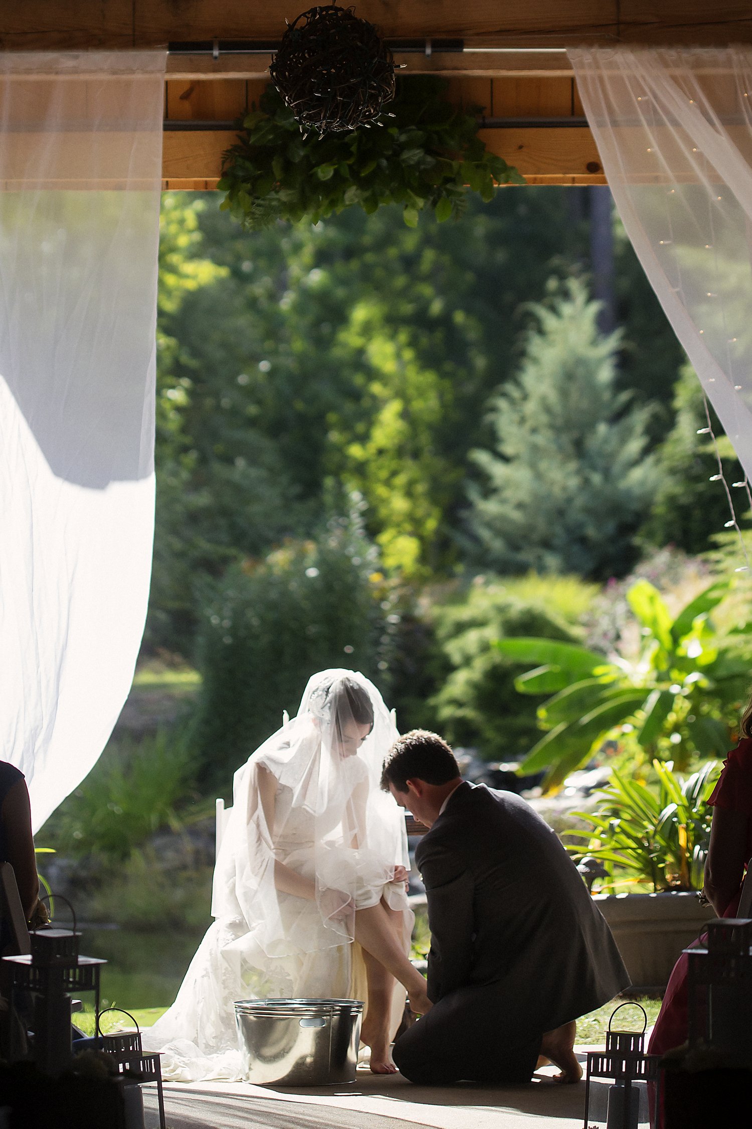 A bride and groom participating in a wedding ceremony outdoors, with the groom kneeling to wash her foot in a silver basin, surrounded by greenery and decorated with sheer white curtains.