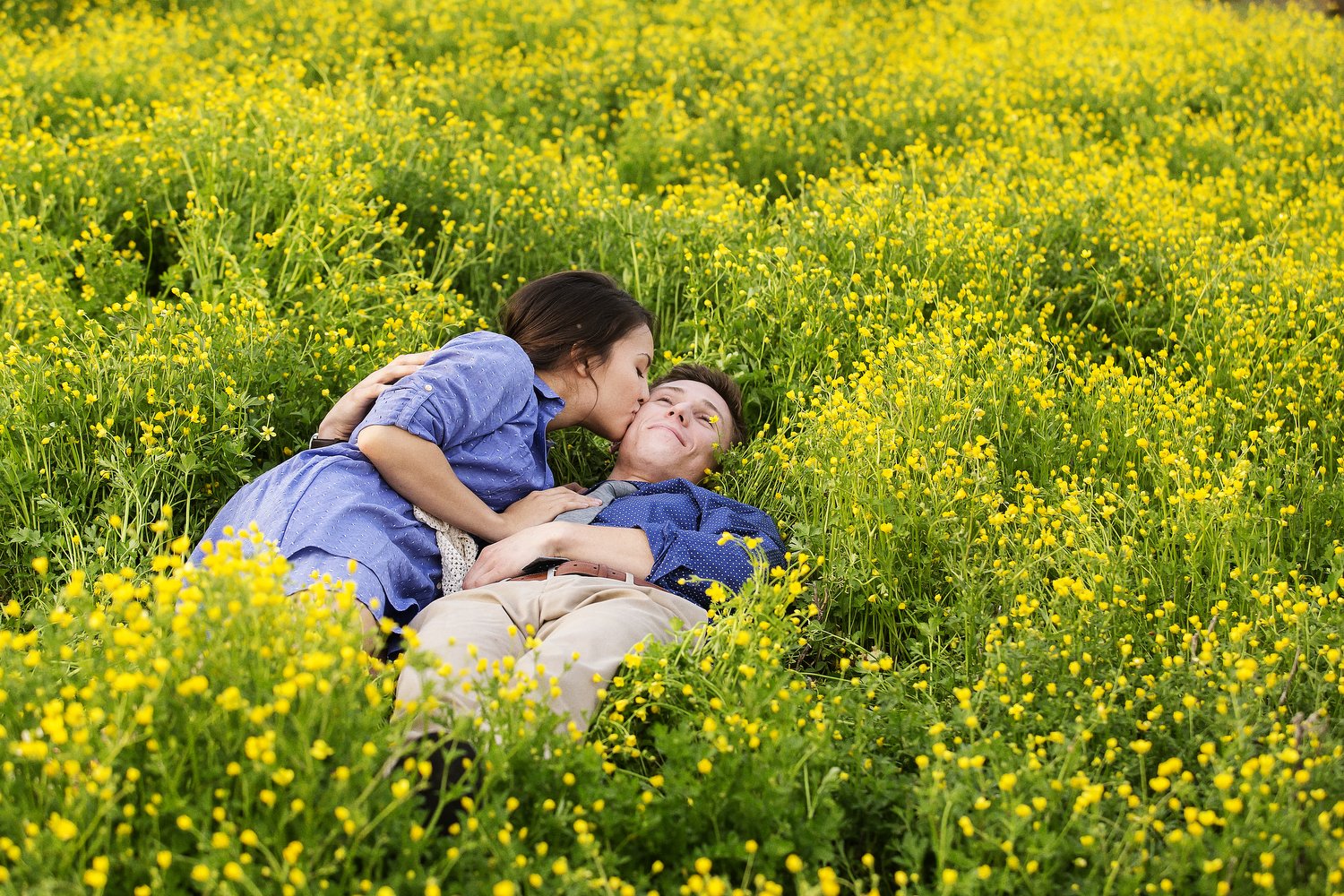 A couple is lying down in a wild field of tiny yellow flowers. You can see an indentation in the field around them. He has on khaki pants and a dark blue dress shirt. She has on a denim dress. He is flat on his back with his right arm behind her as s
