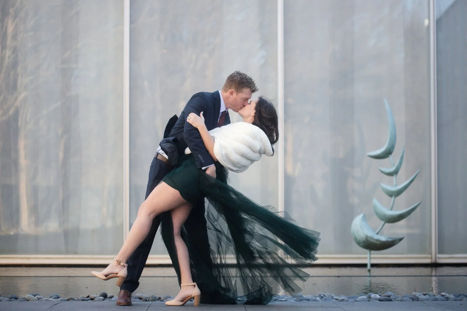 A couple kisses in front of an outdoor museum wall. The walls are glass with white curtains on the inside. There is a teal metal sculpture to the right of the couple. He is wearing a suit and has his arms around her dipping her back to kiss her. She 