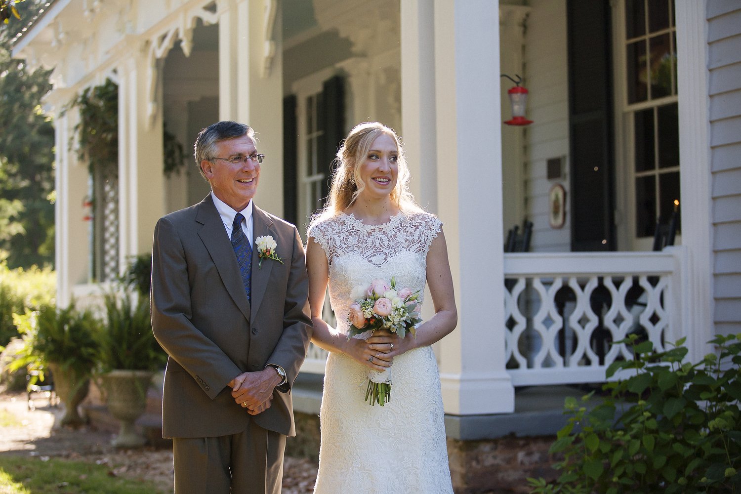 A bride holding a bouquet of pink and white flowers standing next to an older man in a suit outside a house with white columns and a porch, smiling.