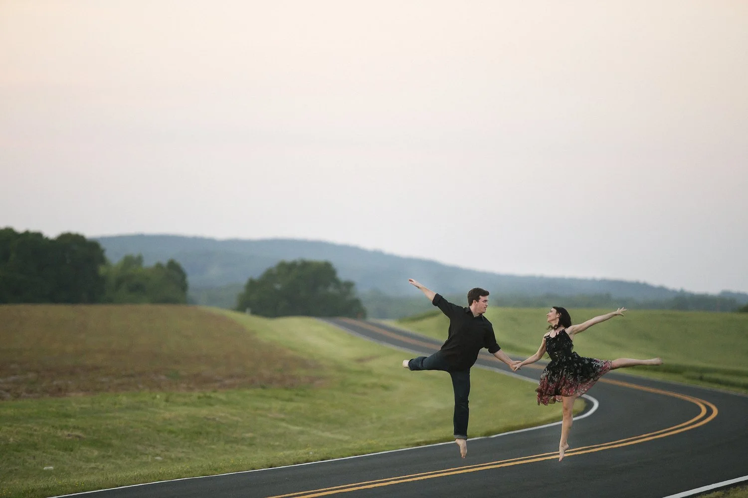 Engagement session with dancers in the street surrounded by grassy fields. They are small in the frame. He has on a black dress shirt and black pants and appears to be either barefoot or wearing points shoes. She has on a black lacy ballerina dress w