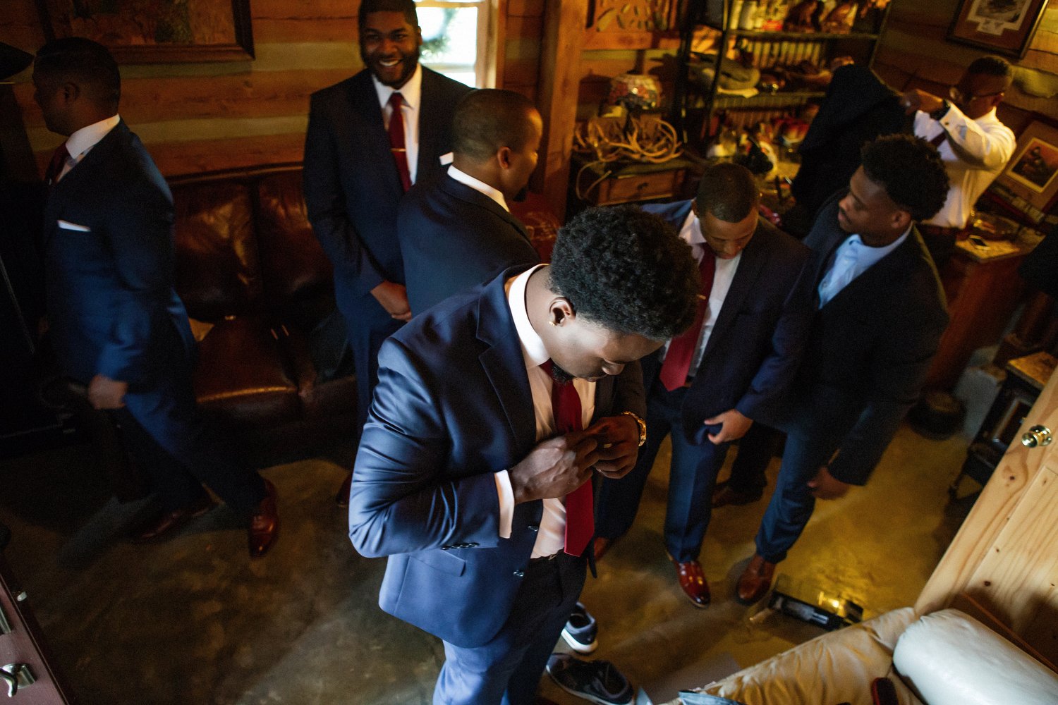 Group of men in navy blue suits with red ties smiling and engaged in conversation. The man in the foreground is adjusting his tie.