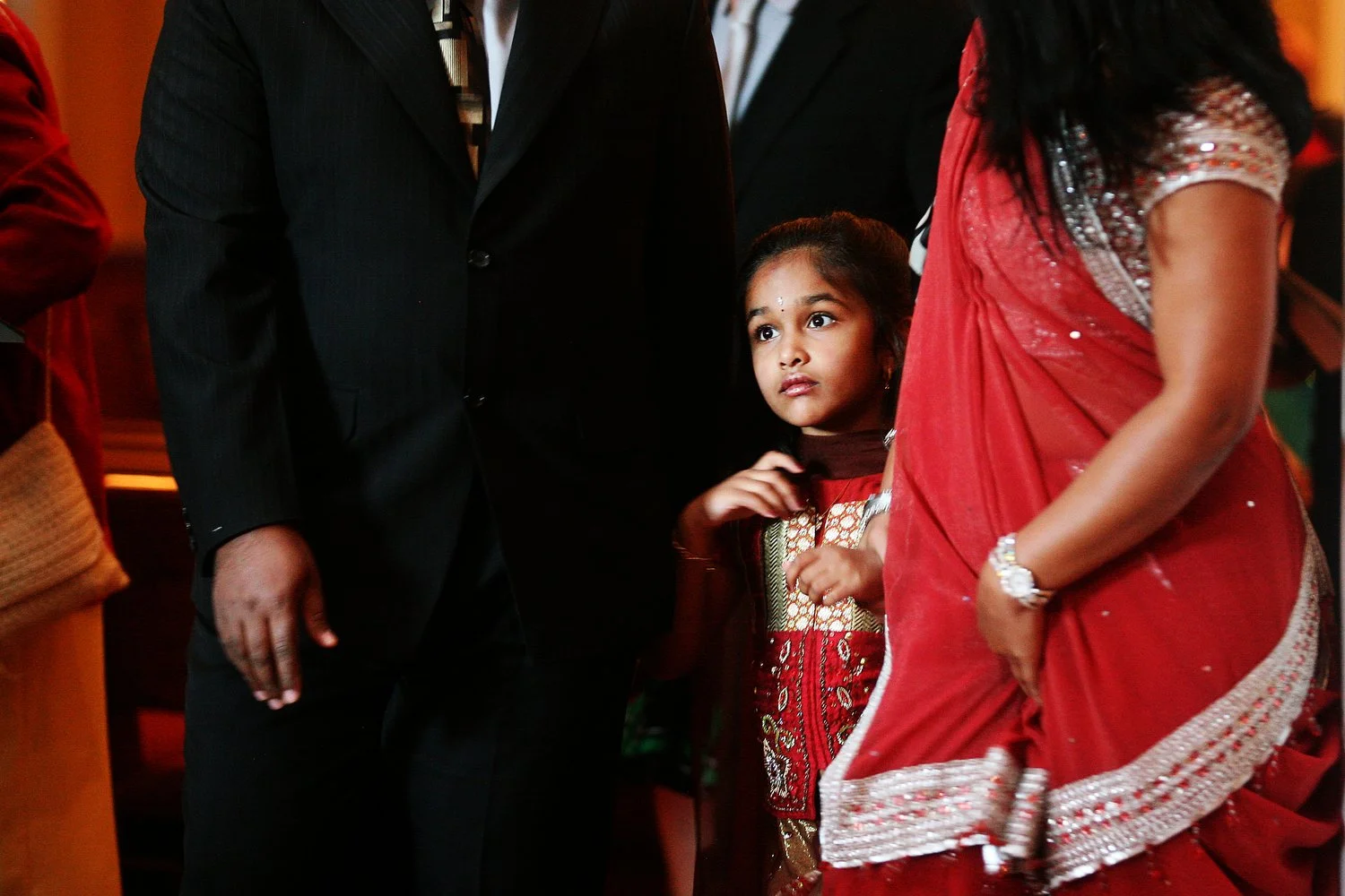 A young girl dressed in red traditional Indian attire standing amid adults wearing formal and traditional dresses at an indoor event.