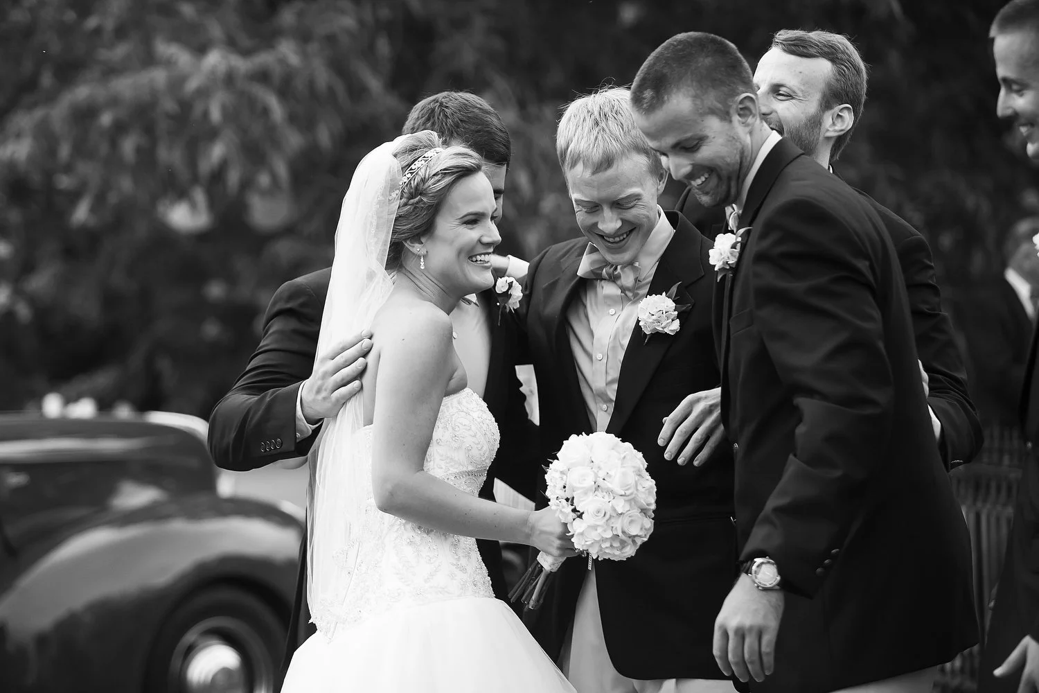 Black and white photo of a bride in a wedding dress with a veil, holding a bouquet of flowers, smiling as she laughs with a group of men dressed in suits, who are also smiling and gathered closely around her.