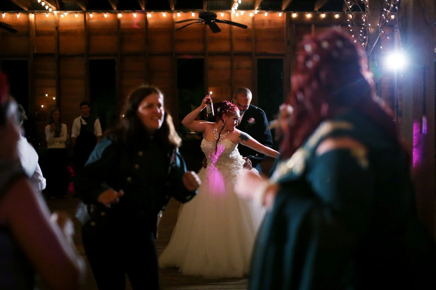 A bride holds a drink above her head as she dances in the distance of a crowded dance floor. She is wearing a white dress with a tulle bottom. In the foreground guests are seen with Renaissance-type clothing. DJ lights from the right beam a white and