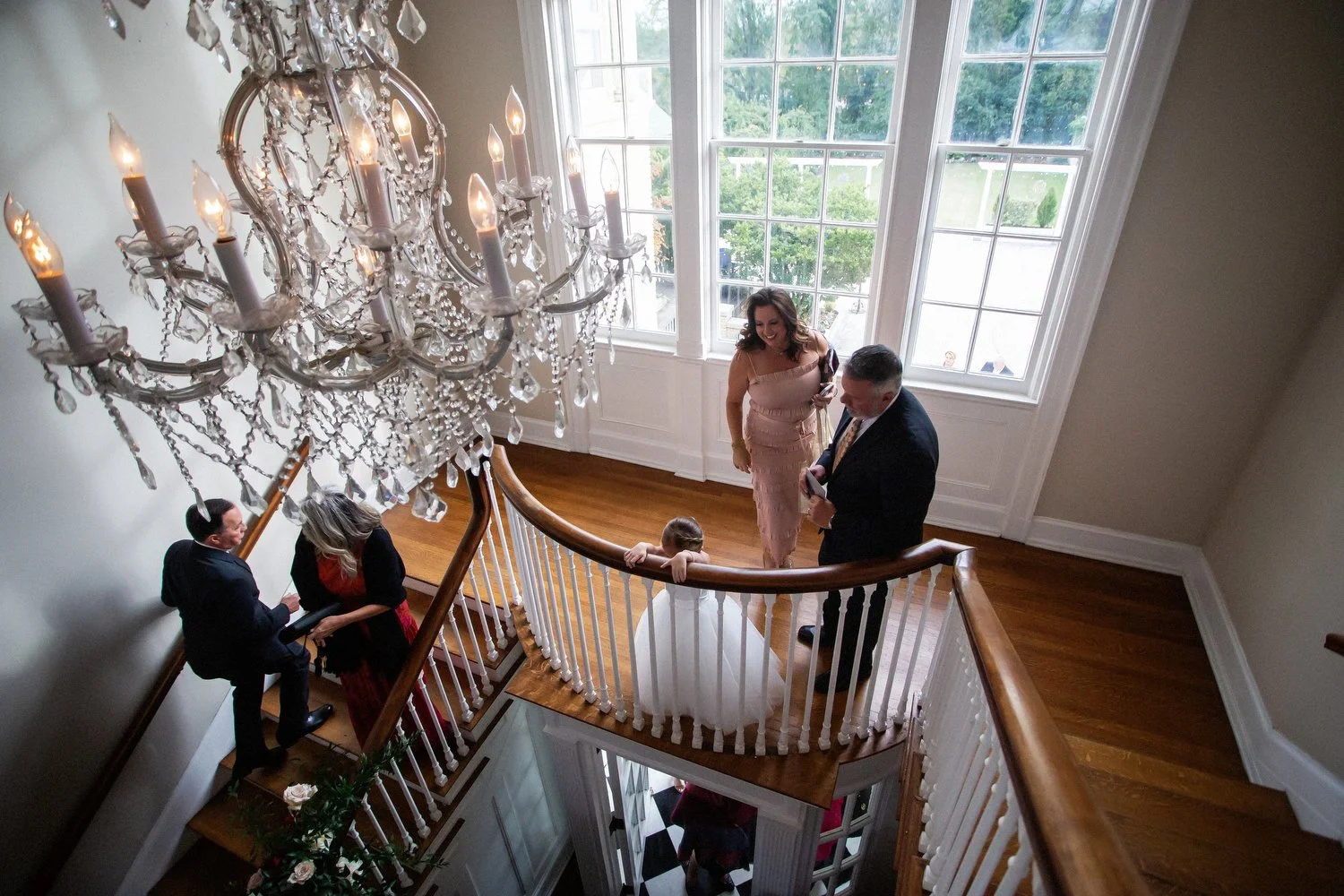 View of a family gathered on a staircase landing before a formal wedding. A chandelier hangs overhead, and large windows let in natural light, showing greenery outside. A woman in a pink dress, a man in a suit, and a child in white tulle are in the c